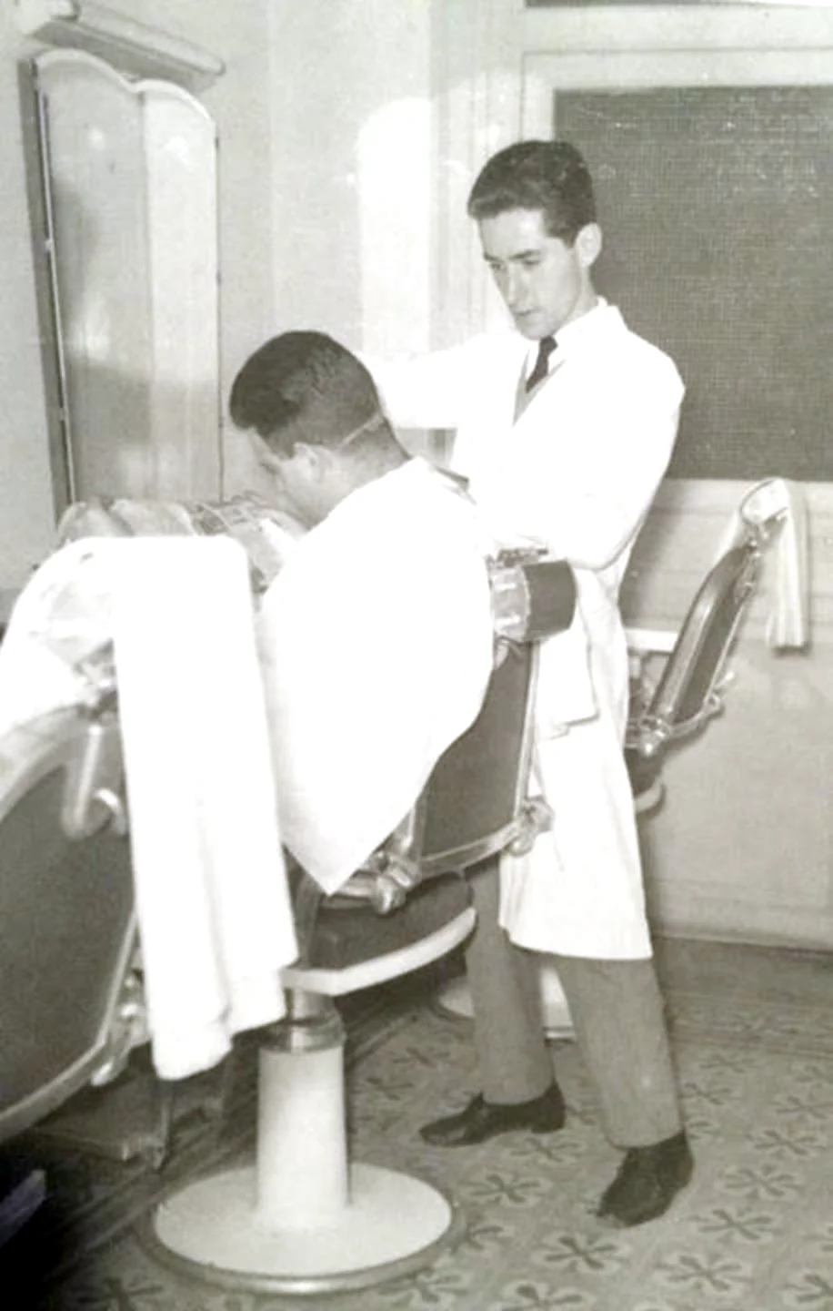 A young barber in a white coat and tie cutting a client's hair at Madison Men's Hair Stylist